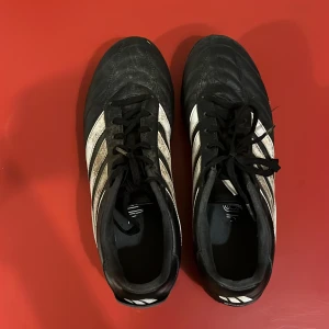 Adidas Gloro svarta fotbollsskor - Säljer ett par klassiska Adidas Gloro fotbollsskor i svart med vita detaljer och de ikoniska tre ränderna på sidorna. Skorna har snörning och är tillverkade i skinn med dobbar för gräsplan. Perfekta för dig som vill ha stilrena och bekväma fotbollsskor.