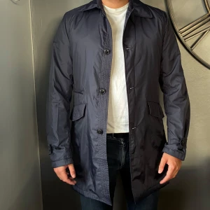 Marinblå trenchcoat J.Lindeberg - Snygg marinblå rock från J.Lindeberg i storlek S. Jackan har klassisk krage, knäppning med knappar, två fickor med lock och bälte bak. Materialet är glansig polyester och insidan har ett diskret mönster. Perfekt för dig som gillar stilrena och moderna plagg. Storleken är någonstans mellan S och M
