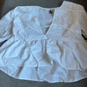 Vit omlottblus med knytband - Säljer en luftig vit omlottblus från H&M med v-ringning och långa ärmar. Blusen har knytband i midjan och en volangig nederdel som ger en avslappnad och trendig look. Perfekt att styla med jeans eller kjol.