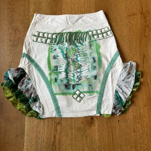 Unik kjol från LeGatte jeans i vitt/grönt - Supercool kjol Legatte jeans strl 38, 55cm lång. Vit med snygga detaljer i olika nyanser grönt, tunna gröna linjer, rundad slits med volang på bägge sidor, påsydda fyrkantiga pärlor på fram och baksida, bred linning upptill, dragkedja i sidan. Väldigt fin