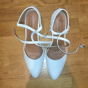 Vita pumps med remmar från Timesize - Snygga vita pumps med spetsig tå och smala remmar som korsas över foten och spänns vid ankeln. Skorna har en stadig klack och är tillverkade i syntetmaterial. Perfekta för dig som vill ha en stilren och trendig look.