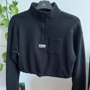 Svart croppad tröja från Adidas - Säljer en svart croppad tröja från Adidas med en halv dragkedja och en liten bröstficka. Tröjan har en avslappnad passform och är perfekt för en sportig look. Den är långärmad och har en elastisk nederkant för extra komfort.