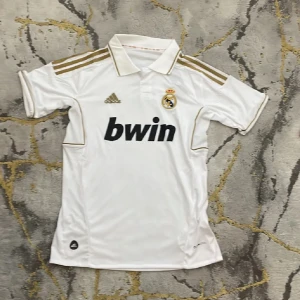 Vit Real Madrid fotbollströja från Adidas - Säljer en snygg vit Real Madrid fotbollströja från Adidas. Tröjan har korta ärmar och är prydd med klubbens emblem på bröstet samt Adidas-loggan. Den har också guldiga detaljer på axlarna och runt kragen. Perfekt för alla Real Madrid-fans!