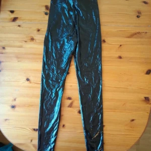 Svarta glansiga leggings - Säljer ett par svarta glansiga leggings som är perfekta för en utekväll eller fest. De är högmidjade och har en tight passform som framhäver benen. Materialet är syntetiskt och ger en cool, glansig look. Perfekta för att sticka ut i mängden!
