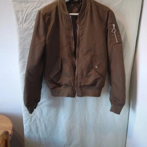 Brun bomberjacka - Säljer en snygg brun/khaki bomberjacka med dragkedja och ribbade muddar. Jackan har en cool ficka med dragkedja på ärmen och två fickor med knappar framtill. Perfekt för höst och vår! Aldrig använd.