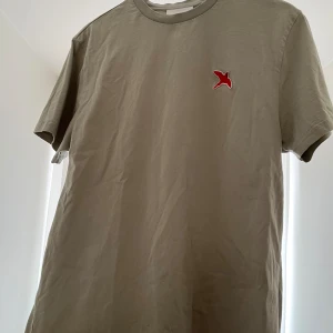Grå t-shirt från Axel Arigato - Grön t-shirt med rött märke från Axel Arigato. använd få gånger, är som nyskick. Något oversized storlek. 