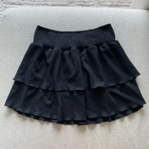 Svart volangkjol - Svart kjol med volanger och töjbar resår i midjan från Gina Tricot. Tillverkad av 98% polyester och 2% elastan. I nyskick då den bara är använd någon enstaka gång.