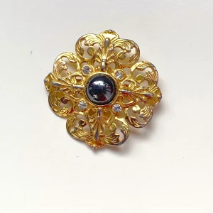 Guldfärgad brosch med svart sten - Säljer en elegant guldfärgad brosch med en stor svart sten i mitten. Troligen hematit. Broschen har ett intrikat mönster med små klara stenar runt om. Perfekt för att ge en touch av glamour till vilken outfit som helst! Okänt material 