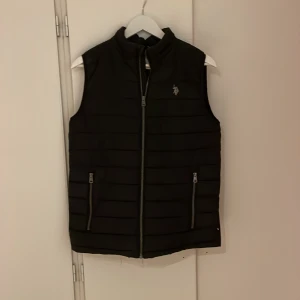 US Polo Assn Väst - En snygg svart Polo Assn väst. Den är fin och stilig, rätt så modern & skön. Bra nu till hösten. Säljer den för att jag använder inte den längre. Den är väldigt använd men har inte så många skador förutom ett litet hål på nedre baksida och få trådar.