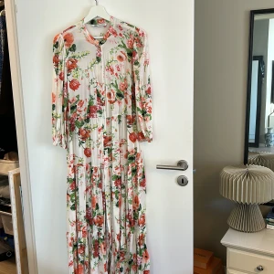 Blommig långklänning från Zara - Säljer en superfin blommig långklänning från Zara. Den har ett härligt mönster med röda och gröna blommor på en vit bakgrund. Klänningen har långa ärmar och en avslappnad passform, perfekt för vår och sommar. Den är gjord i ett lätt och luftigt material, vilket gör den idealisk för varmare dagar. Perfekt för både vardag och festliga tillfällen!