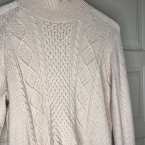 Beige stickad tröja från Bubbleroom - Säljer en snygg beige stickad tröja från Bubbleroom. Tröjan har ett detaljerat flätmönster på framsidan och en hög krage. Perfekt för kyliga dagar!
