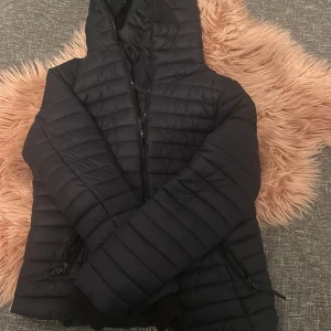 Svart pufferjacka från Marikoo - Snygg svart pufferjacka från Marikoo med huva och dragkedja. Jackan har en quiltad design och praktiska fickor med dragkedjor. Perfekt för kyligare dagar.