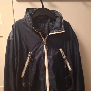 Mörkblå vindjacka med dragkedjor (Armani) - Snygg mörkblå vindjacka med vita dragkedjor och hög krage. Perfekt för blåsiga dagar och har praktiska fickor med dragkedja. Enkel och stilren design som passar till det mesta.