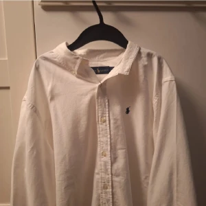 Vit skjorta från Ralph Lauren - Klassisk vit skjorta från Ralph Lauren med broderad logga på bröstet. Skjortan har långa ärmar och knappar framtill. Perfekt för en stilren look.