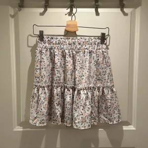 Blommig kortkjol med volang - Charmig kortkjol i vitt med ett färgglatt blommönster. Kjolen har en elastisk midja och volangdetaljer som ger en lekfull look.