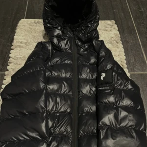 Svart pufferjacka från Peak Performance - Snygg svart pufferjacka från Peak Performance med glansig yta och huva. Jackan har en dragkedja framtill och är perfekt för kyliga dagar. Den är designad för att hålla dig varm och bekväm. 