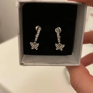 Silverfärgade fjärilsörhängen med stenar - Fick dessa som födelsedagspresent, men har aldrig använt, inte ens testat. Helt nyskick alltså. Eleganta silverfärgade örhängen med gnistrande stenar. De har en hängande design med en liten fjärilsformad detalj längst ner. Perfekta för att ge en glittrig touch till din outfit. Lådan medföljes :)