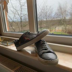 Svarta sneakers med broderad detalj - Snygga svarta sneakers med broderade detaljer i rött och orange på sidorna. Skorna har en klassisk snörning och en vit sula som ger en stilren look. Perfekta för en avslappnad stil.