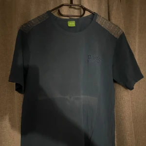 Blå t-shirt från Hugo Boss - Snygg blå t-shirt från Hugo Boss med broderad logga på bröstet. T-shirten har korta ärmar och ett stilrent mönster på axlarna med texten 'HUGO BOSS' i grönt. Perfekt för en casual look.