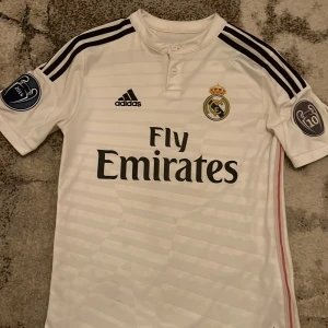 Vit Real Madrid fotbollströja - Säljer en vit Real Madrid fotbollströja från Adidas med korta ärmar. Tröjan har svarta detaljer och klubbens emblem på bröstet. Den har även 'Fly Emirates' som sponsorlogga och 'Ronaldo 7' på ryggen. Tröjan är helt äkta och finns inte att köpa längre i butiker
