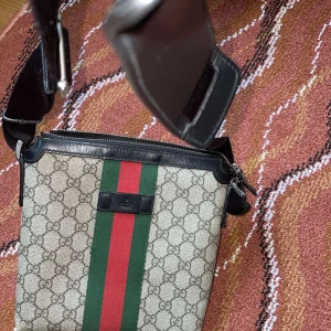 Beige axelväska med ränder - Snygg beige axelväska med klassiskt mönster och gröna samt röda ränder. Väskan har en justerbar axelrem och dragkedja upptill. Perfekt för att bära dina nödvändigheter med stil.