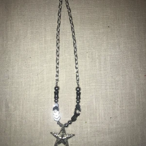 Silverfärgat halsband med stjärnhänge - Unikt halsband med ett stort silverfärgat stjärnhänge. Kedjan är dekorerad med svarta och silverfärgade pärlor samt små vita pärlor. Perfekt för att ge en glittrig touch till din outfit.