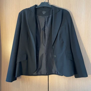 Svart kavaj från Kookai - Elegant svart kavaj från Kookai med klassisk design. Kavajen har två knappar framtill och är tillverkad i ett stilrent material som ger en sofistikerad look. Perfekt för både formella och semi-formella tillfällen.