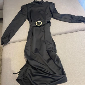 Svart klänning från Twist & Tango - Elegant svart klänning från Twist & Tango med långa ärmar och en stilren design. Klänningen har en dekorativ knytning i sidan för en snygg siluett. Perfekt för en chic look. Bälte medföljer ej.