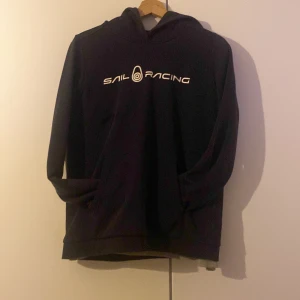 Mörkblå hoodie från Sail Racing - Säljer en mörkblå hoodie från Sail Racing med vit logga på bröstet. Tröjan har en klassisk design med huva och långa ärmar. Perfekt för en avslappnad stil, den är i jätte fint skick och varm med skönt matreal inuti.