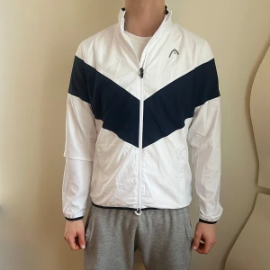 Vit och marinblå vindjacka från Head - Säljer en ny stilren vit och marinblå tennis/padel vindjacka från Head. Jackan har en dragkedja framtill och ett sportigt mönster med en mörkblå V-formad detalj över bröstet. Perfekt för träning eller en aktiv vardag. Storlek XL dam men passar tight på mig vid axel/armparti som är herr 185cm lång smal dryga 80kg. Nypris 830kr