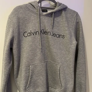 Grå hoodie från Calvin Klein - Säljer en stilren grå hoodie från Calvin Klein Jeans med svart logotyp på bröstet. Tröjan har en klassisk känguruficka och justerbar huva med dragsko. Perfekt för en avslappnad look.