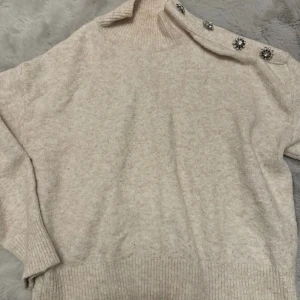 Beige stickad tröja med dekorativa knappar - Mysig beige stickad tröja med unika dekorativa knappar på axeln. Tröjan har en hög krage och långa ärmar, perfekt för kyligare dagar. Den ribbade kanten ger en snygg finish.