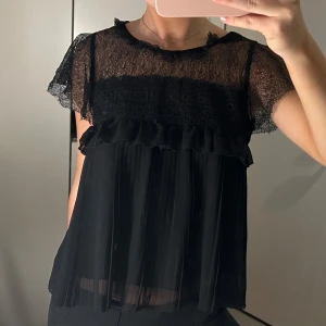 Svart spetsblus med volangdetaljer från Zara - Elegant svart blus i spets med korta ärmar och volangdetaljer. Perfekt för en stilfull look med transparenta partier och vacker struktur.