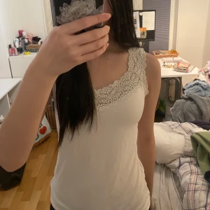 Vit ärmlös topp med spetsdetaljer - Säljer en söt vit ärmlös topp med vackra spetsdetaljer runt halsringningen. Perfekt för varma sommardagar eller som ett lager under en kofta. Toppen har en enkel och stilren design.