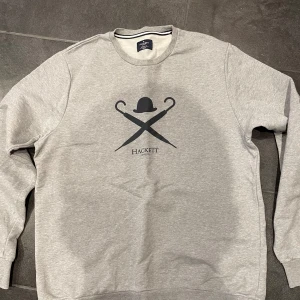 Grå sweatshirt från Hackett - Säljer en stilren grå sweatshirt från Hackett med ett klassiskt tryck av en hatt och korsade paraplyer på framsidan. Tröjan har rund hals och långa ärmar, perfekt för en avslappnad stil.