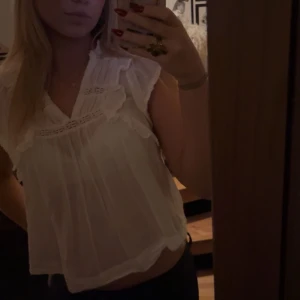 Vit blus med spetsdetaljer - Säljer en söt vit blus med spetsdetaljer vid halsen. Blusen har korta ärmar och en luftig passform, perfekt för varmare dagar. Den är lätt och bekväm att bära.