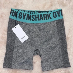 Grå träningsshorts från Gymshark - Snygga grå träningsshorts från Gymshark med en bred, turkos resårmidja och svart text. Perfekta för gymmet eller löprundan. De är tillverkade i ett stretchigt material som ger en tight passform. Alldrig använda!