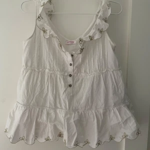 Vit ärmlös blus från Orsay - Charmig vit blus från Orsay med volangdetaljer och knappar framtill. Blusen har broderade kanter med små rosetter och en luftig passform, perfekt för varma dagar.