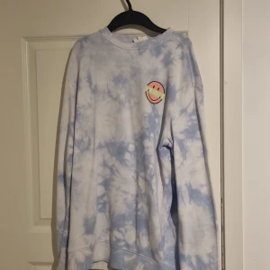 Blå och vit tie-dye tröja med smiley-motiv - Säljer en cool blå och vit tie-dye tröja med flera smiley-motiv. Tröjan har långa ärmar och en avslappnad passform. Perfekt för en avslappnad stil.