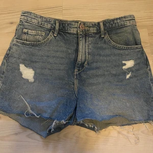 Blå jeansshorts med slitningar - Säljer ett par coola blå jeansshorts med slitna detaljer och fransig kant. Shortsen har en klassisk femficksdesign och stängs med dragkedja och knapp. Perfekta för en avslappnad stil! Pris går att diskutera💗