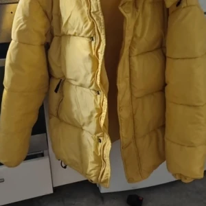 Gul pufferjacka från Gant - Säljer en snygg gul pufferjacka från Gant. Jackan har en quiltad design med dragkedja och knappar framtill. Perfekt för kyligare dagar med sin varma och bekväma passform.