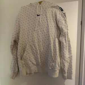 Vit hoodie från Nike med mönster - Säljer en vit hoodie från Nike med ett diskret mönster och svart logga på bröstet. Hoodien har en justerbar huva med snören och en stor ficka framtill. Ärmarna har en svart Nike-remsa längs sidan. Perfekt för en sportig look!