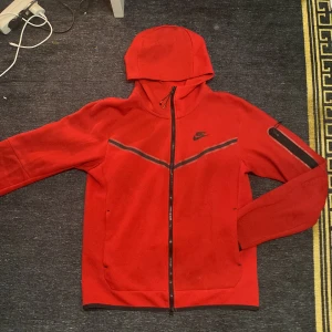 Röd hoodie Nike tech fleece  - Säljer en röd hoodie från Nike med dragkedja och huva. Jackan har svarta detaljer och en ficka med dragkedja på ärmen. Perfekt för en sportig look.