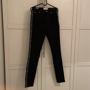 Svarta jeans från Calvin Klein - Snygga svarta jeans från Calvin Klein med logotyp längs sidorna. De har en klassisk femficksdesign och knappgylf. Perfekta för en stilren look.