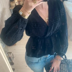 Svart omlottblus med spetsdetaljer - Snygg svart omlottblus med brodyr detaljer och långa ärmar. Perfekt för en stilren look med v-ringad design och elegant passform.