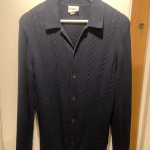 Mörkblå cardigan/kofta med knappar - Snygg mörkblå kofta med kabelstickat mönster och knappar framtill. Perfekt för kyligare dagar och ger en stilren look. Långärmad design med klassisk krage. Pris går att diskutera!