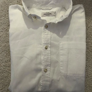 Vit linneskjorta från Dressmann - Klassisk vit skjorta från Dressmann med knappar och bröstficka. Perfekt för en stilren look.