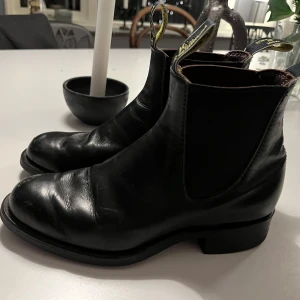 Svarta boots från R.M. Williams - Snygga svarta boots i skinn från R.M. Williams. Klassisk design med elastiska paneler på sidorna och dragflikar för enkel påtagning. Perfekta för både vardag och mer uppklädda tillfällen.