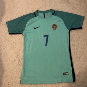  Portugal 2016 Em Ronaldo  - Säljer en grön Portugal fotbollströja från Nike med nummer 7 och namn på ryggen. Tröjan har korta ärmar och är i Dri-FIT material för optimal komfort. Perfekt för fotbollsfans!
