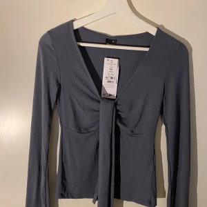 Grå långärmad topp med knyt - Snygg grå långärmad topp med knyt framtill. Perfekt för en stilren look. Toppen har en djup v-ringning och är tillverkad i ett mjukt material.